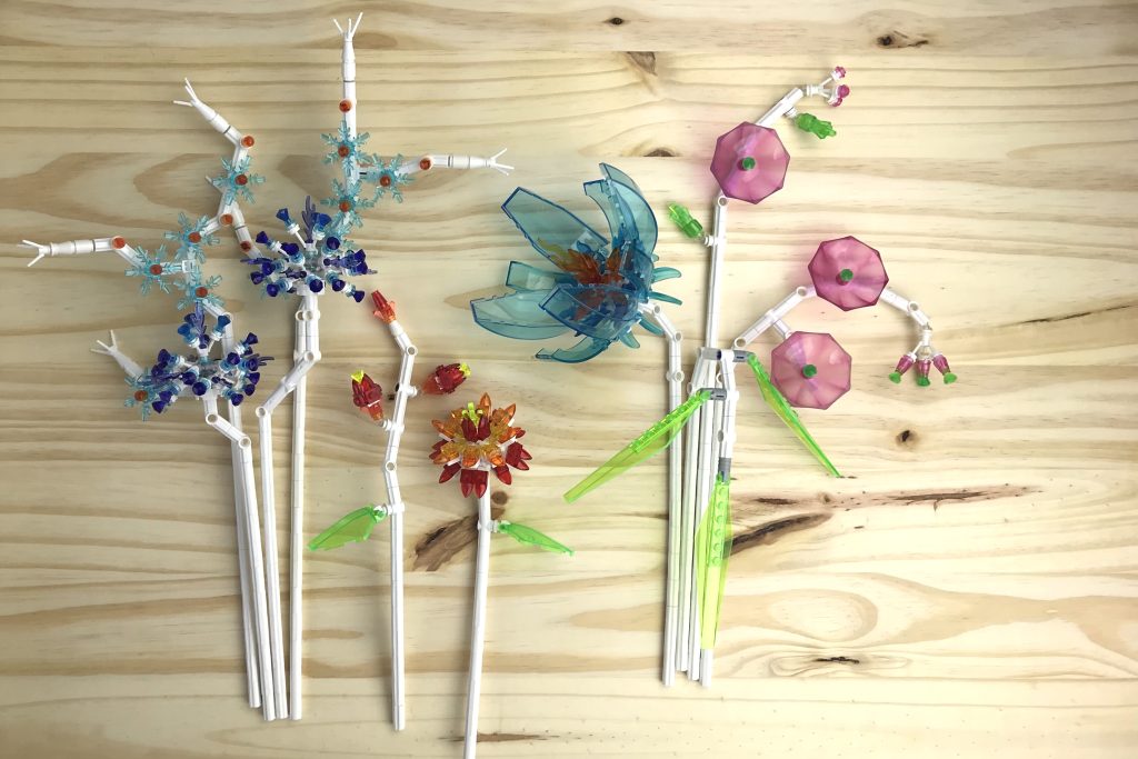 Lego brick-built flowers made from transparent parts and white stems lie on a pine board. Transparent pieces include light blue snowflakes, dark purple gems, red and orange tooth plates, bright green flaps with fork, light blue and bright green cockpits, and pink umbrellas.