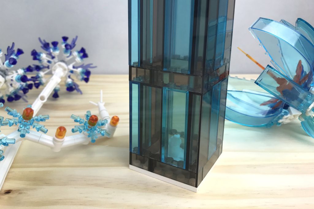 A closeup of a LEGO brick-built transparent base with a white "flower frog" pin inside (built from an actual LEGO frog) for floral arranging. LEGO flowers can be seen next to the vase on the table, including a light blue cockpit flower, a dark purple gem flower, and a light blue snowflake flower.