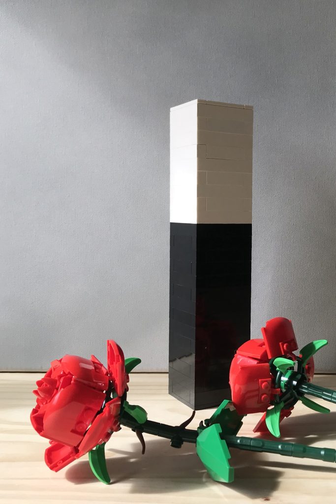 A tall, narrow rectangular simple colorblock brick-built bud vase sits on a wooden surface next to two red LEGO roses, against a light gray background.