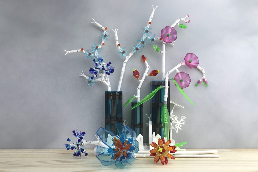 LEGO brick-built flowers made from transparent blue, purple, red, orange, pink, and green parts and white stems are arranged in three vases. The vases sit in a snowy landscape with a white tree. Three flowers of different shapes and sizes lie on the wooden table next to the base.