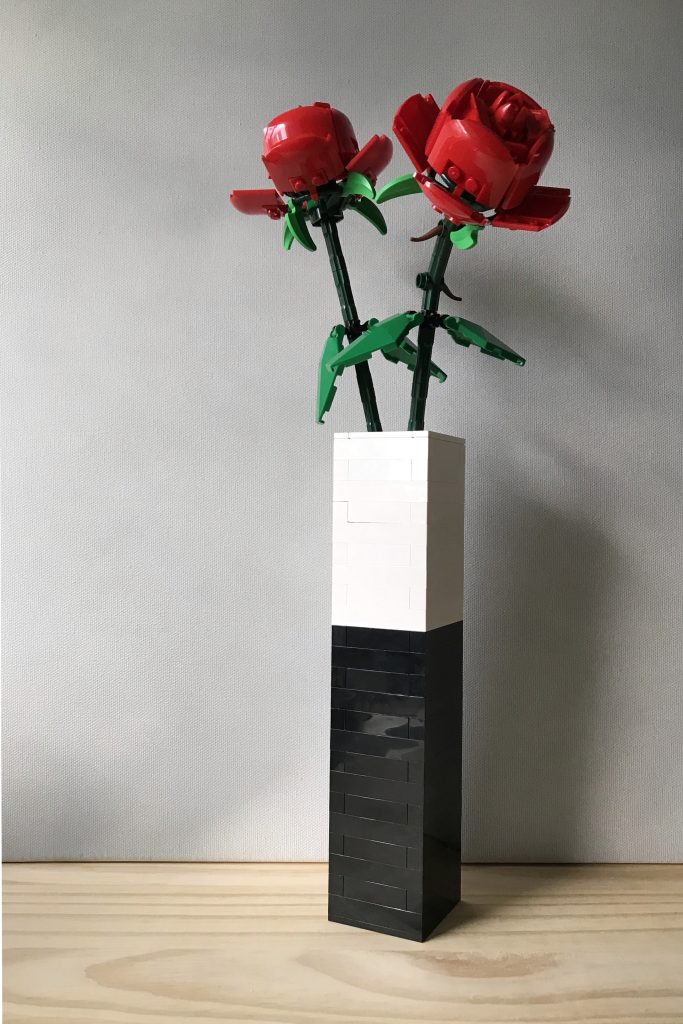 A tall, narrow rectangular simple black and white colorblock brick-built bud vase holding two red LEGO roses sits on a wooden surface, against a light gray background.