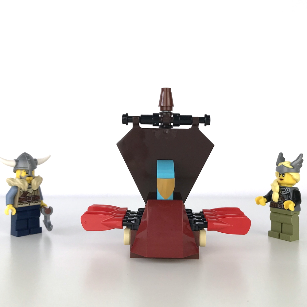 Mini version of a LEGO Viking Ship, with a brown sail and red oars, front view. Two Viking minifigs examine it skeptically, as it's too small for them to ride in.