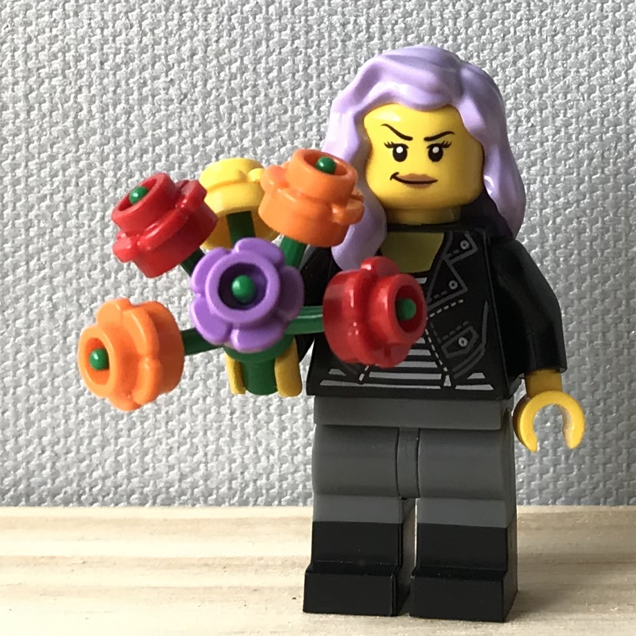 A smirking LEGO minifig with classic yellow skin and long lavender hair holds a plant stem with six colorful five-petalled flowers. She is wearing a striped shirt, black leather jacket, gray pants, and black boots. She stands on a light wood floor in front of a light gray canvas wall.