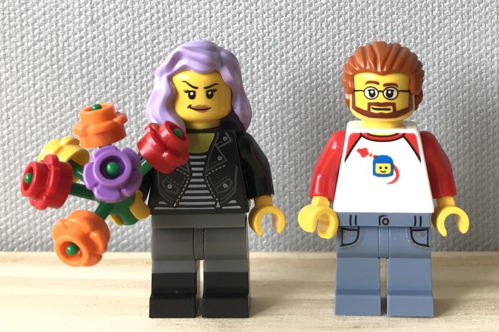 Two classic yellow LEGO minifigs stand together: a woman with lavender hair and a man with glasses and a dark orange mustache, goatee, and hair. The woman holds a bouquet of flowers. The man wears a classic space helmet shirt.