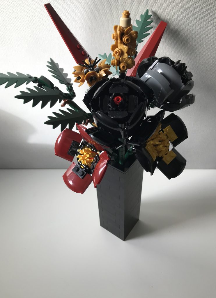 Simple black LEGO vase with LEGO flowers: red and black poppies, black and gray roses, pearl gold lavender and cornflowers, dark green ferns, and dark red leaves, on a white desk against a light gray background.