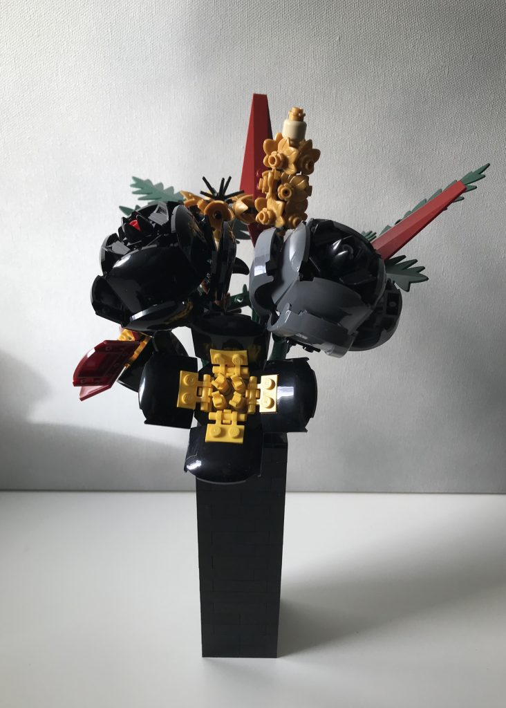 Simple black LEGO vase with LEGO flowers: red and black poppies, black and gray roses, pearl gold lavender and cornflowers, dark green ferns, and dark red leaves, on a white desk against a light gray background.