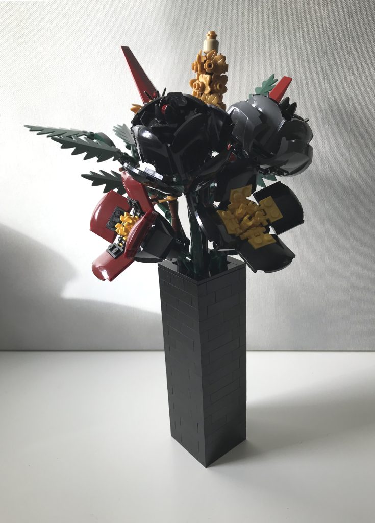 Simple black LEGO vase with LEGO flowers: red and black poppies, black and gray roses, pearl gold lavender and cornflowers, dark green ferns, and dark red leaves, on a white desk against a light gray background.