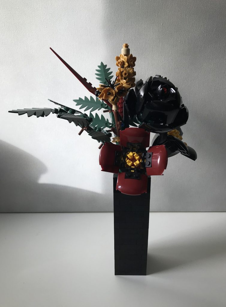 Simple black LEGO vase with LEGO flowers: red and black poppies, black and gray roses, pearl gold lavender and cornflowers, dark green ferns, and dark red leaves, on a white desk against a light gray background.