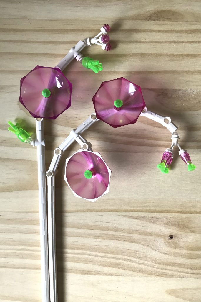 LEGO brick-built flowers MOC with white technic stems, transparent dark pink umbrellas as flowers, with transparent bright green gem centers, along with pink buds and green clusters of new leaves, on a light wood background.