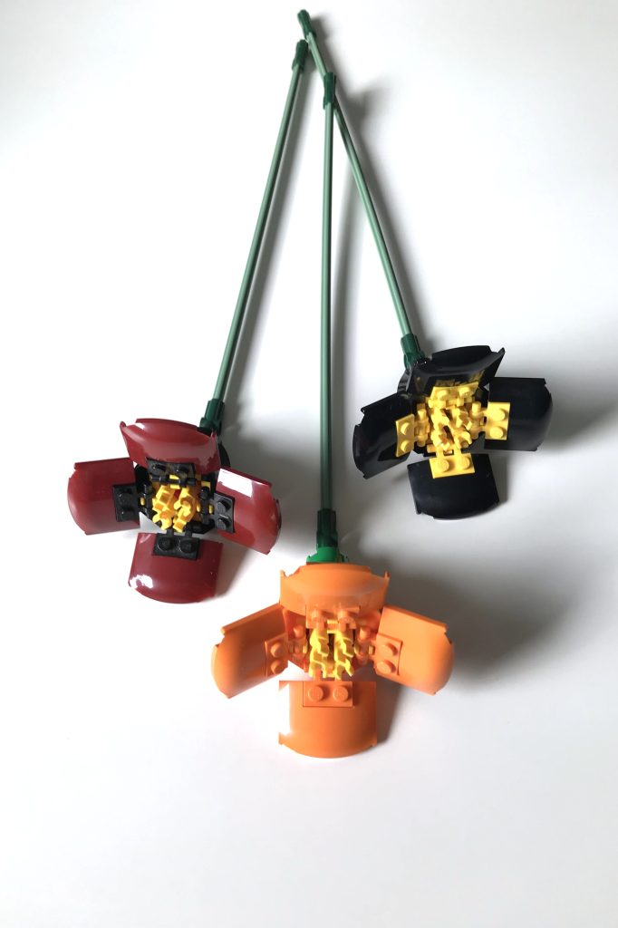 Three LEGO poppy flowers in dark red, orange, and black and yellow lie on a white surface.