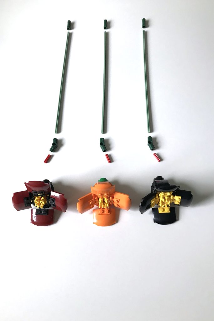 Three LEGO poppy flowers in dark red, orange, and black and yellow lie on a white surface in front of stem pieces.