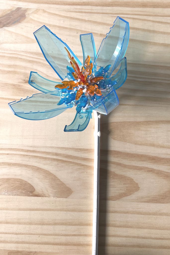 A large LEGO flower with a white stem, transparent light blue cockpit petals, and transparent blue and orange flame center parts, lying on a light wood surface.