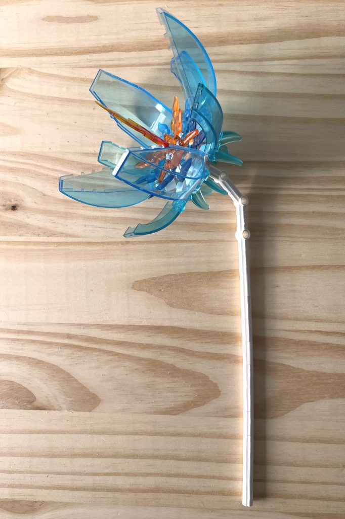 A large LEGO flower with a white stem, transparent light blue cockpit petals, and transparent blue and orange flame center parts, lying on a light wood surface.