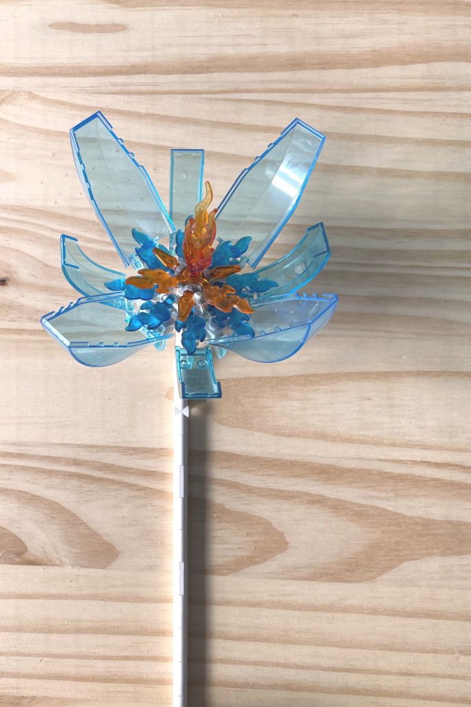 A large LEGO flower with a white stem, transparent light blue cockpit petals, and transparent blue and orange flame center parts, lying on a light wood surface.