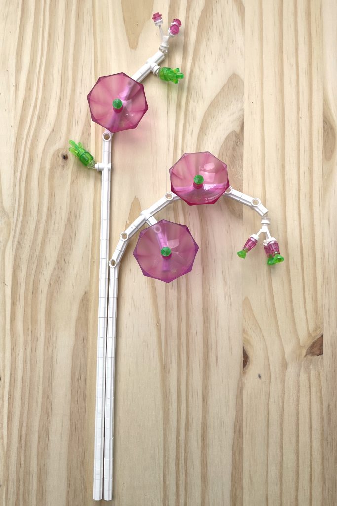 Sprays of LEGO flowers with white stems and transparent pink and bright green umbrella flowers lying on a light wood surface.