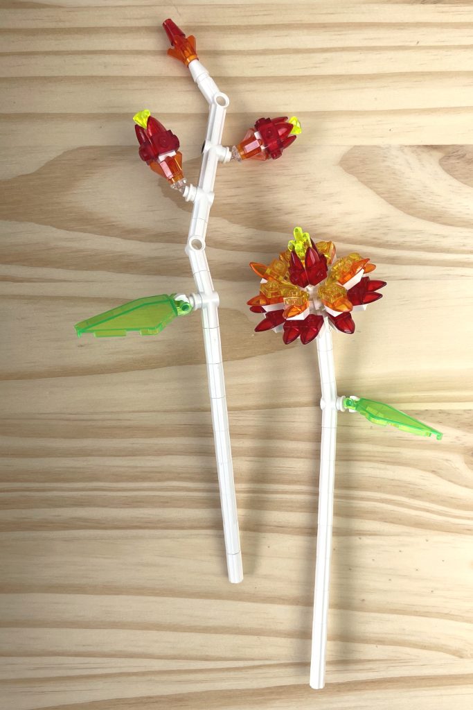 LEGO flowers with white stems, transparent bright green leaves, and transparent orange and red flowers made from tooth plates lying on a light wood surface.