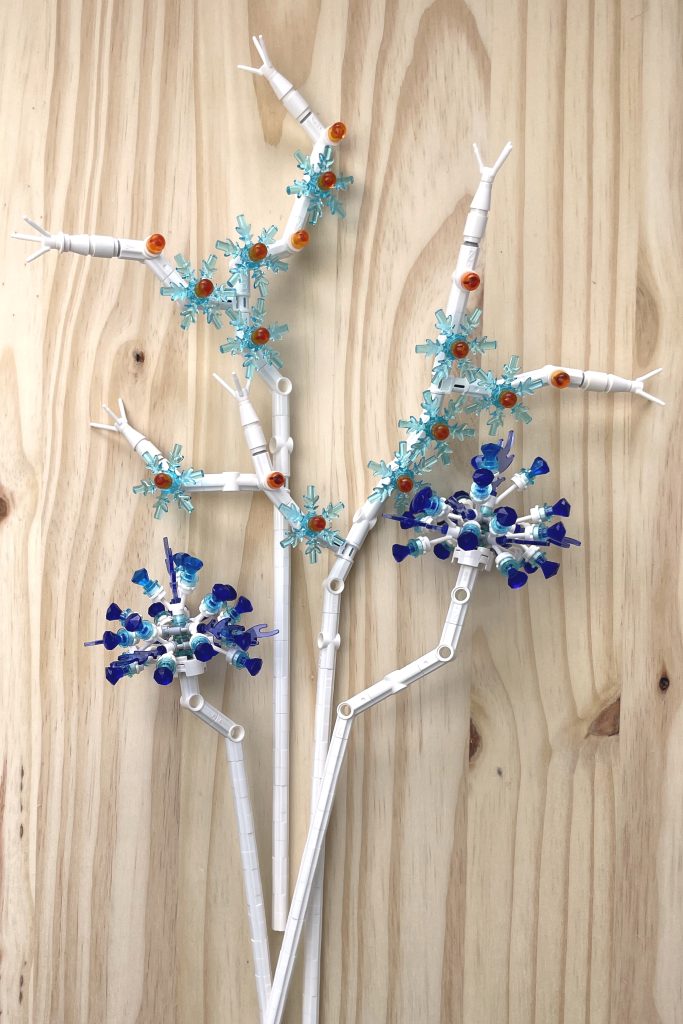 Sphere-shaped allium-like flowers built from white LEGO stems and transparent purple gems and flames and sprays of transparent light blue and orange LEGO snowflake flowers lying on a light wood surface.