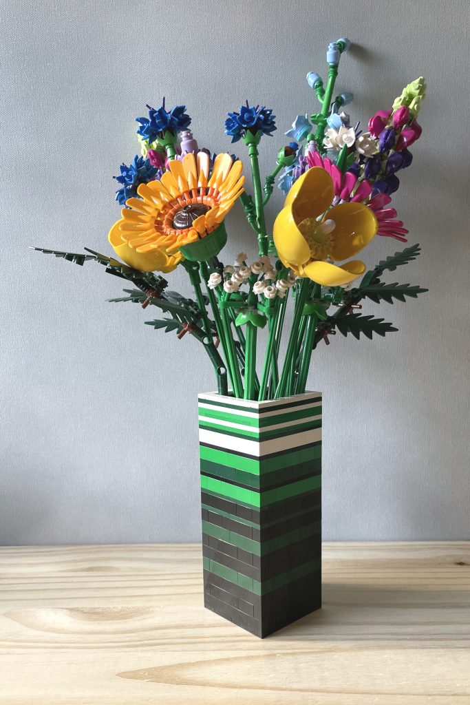 LEGO vase built with bricks and plates in white, green, dark green, and black to for stripes, holding LEGO's Wildflower bouquet, on a light wood surface in front of a light gray background.