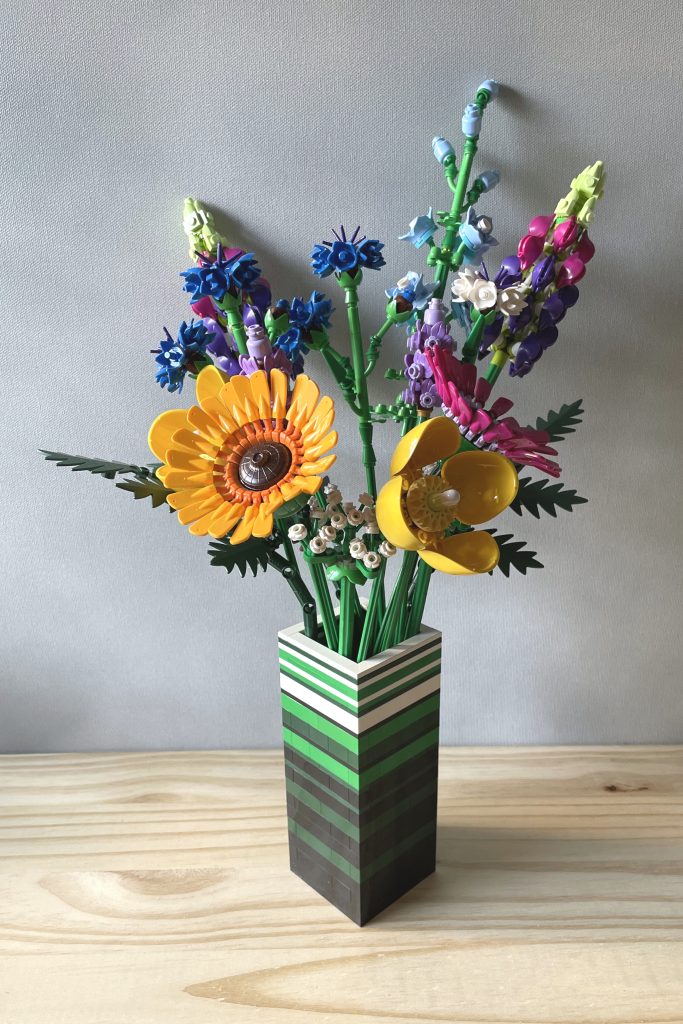 LEGO vase built with bricks and plates in white, green, dark green, and black to for stripes, holding LEGO's Wildflower bouquet, on a light wood surface in front of a light gray background.