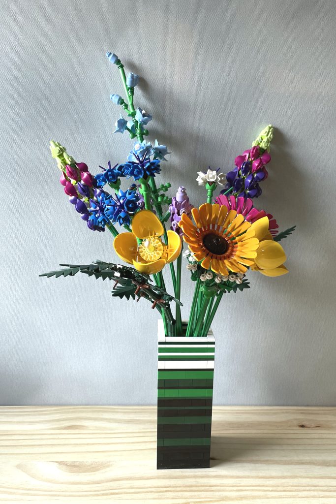 LEGO vase built with bricks and plates in white, green, dark green, and black to for stripes, holding LEGO's Wildflower bouquet, on a light wood surface in front of a light gray background.