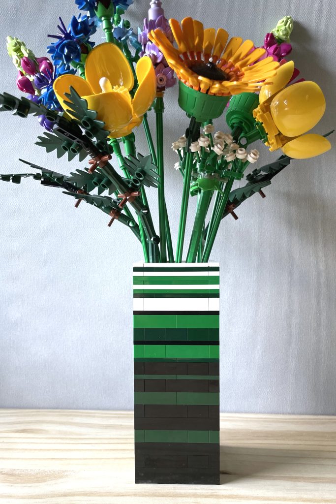 Closeup of a LEGO vase built with bricks and plates in white, green, dark green, and black to for stripes, holding LEGO's Wildflower bouquet, on a light wood surface in front of a light gray background.