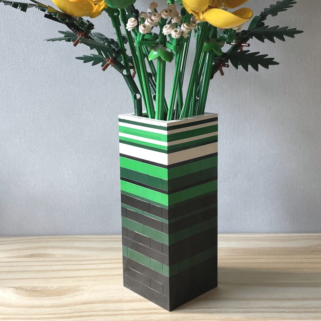 Closeup of a LEGO vase built with bricks and plates in white, green, dark green, and black to for stripes, holding LEGO's Wildflower bouquet, on a light wood surface in front of a light gray background.